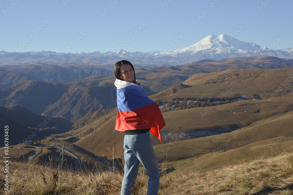 A young brunette woman stands against the backdrop of the snow-capped Mount Elbrus, looking at the camera, a Russian flag covers her shoulders. Tricolor against the backdrop of snow-capped Mount