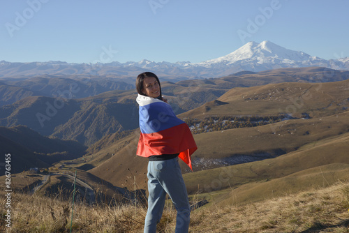 Obraz na płótnie A young brunette woman stands against the backdrop of the snow-capped Mount Elbrus, looking at the camera, a Russian flag covers her shoulders. Tricolor against the backdrop of snow-capped Mount w salonie
