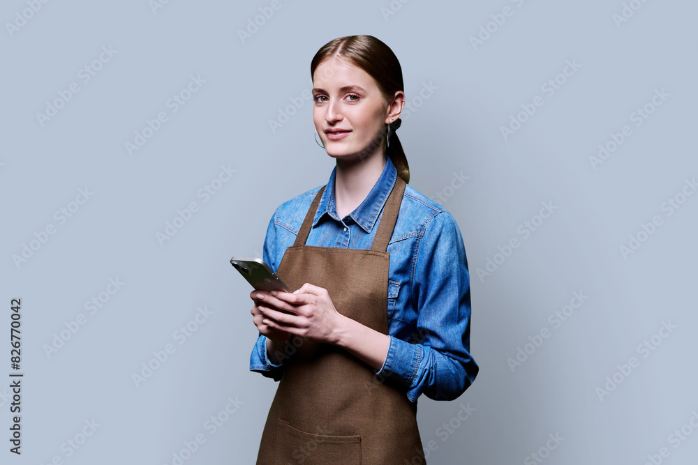 Young female worker in an apron using smartphone on gray studio background Young female worker in an apron using smartphone on gray studio background