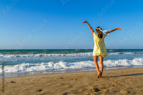 Carefree young happy stress free woman, feeling alive, free in nature with arms rased up, breathing clean fresh air at the beach, standing on wet sand looking at the blue ocean horizon.