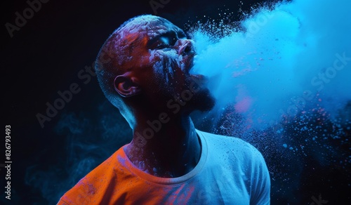 Wide shot of an African American man wearing a neon colored t-shirt  blowing blue powder from his mouth into the air against a black background.