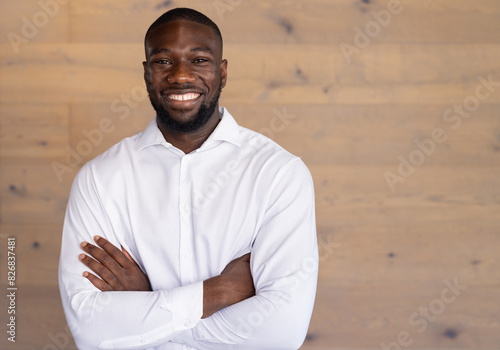In a modern office, a young African American man stands with arms crossed and smiling, copy space photo