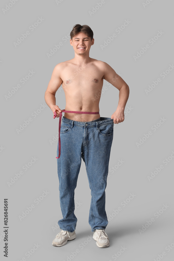Sporty young man in loose jeans and with measuring tape on grey background. Diet concept Sporty young man in loose jeans and with measuring tape on grey background. Diet concept