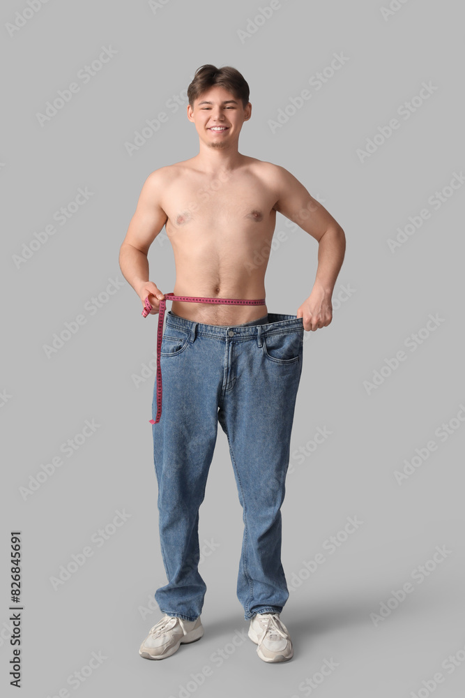 Sporty young man in loose jeans and with measuring tape on grey background. Diet concept