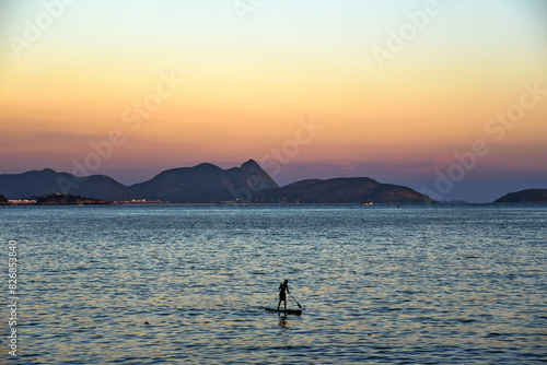 Fotografi A Lone Paddleboarder in the Waters of Praia Vermelha at Dusk - Rio de Janeiro, B