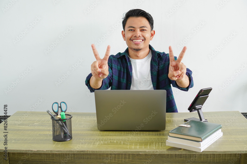asian man sitting in workplace gesturing peace sign with fingers. Happy guy enjoying remote work at home. Concept of freelance and technology. asian man sitting in workplace gesturing peace sign with fingers. Happy guy enjoying remote work at home. Concept of freelance and technology.
