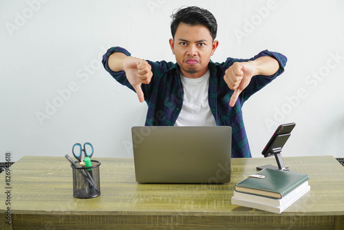 Displeased young asian businessman sitting office workplace showing thumbs down gesture