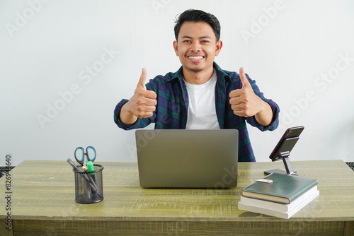 young asian freelancer sitting in workplace doing happy thumbs up gesture with hand. approving expression looking at the camera showing success.