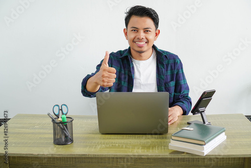 young asian freelancer sitting in workplace doing happy thumbs up gesture with hand. approving expression looking at the camera showing success.