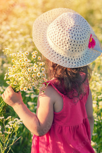 Happy little girl in a straw hat holding a bouquet of daisies and walking through a flower field on a sunny summer day © vvvita