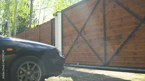 The gate of a private property opens, a black sedan drives in and out of the house on a sunny day. Long version