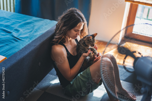Young woman with a gray cat in her arms sitting at home on the floor in front of a fan, escapefrom the summer heat concept