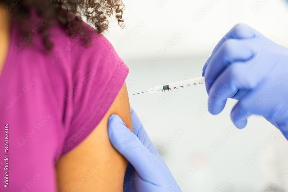 Close-up of a doctor administering a vaccine injection into a patient's arm. The patient is wearing a purple shirt, and the doctor's gloved hands are holding the syringe. Close-up of a doctor administering a vaccine injection into a patient's arm. The patient is wearing a purple shirt, and the doctor's gloved hands are holding the syringe.
