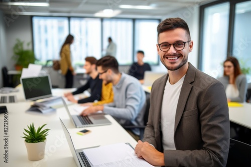 Portrait of a young male IT professional sitting at desk, holding document, smiling and looking at camera at office with his coworkers in background