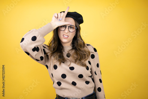 Young beautiful brunette woman wearing french beret and glasses over yellow background making fun of people with fingers on forehead doing loser gesture mocking and insulting.