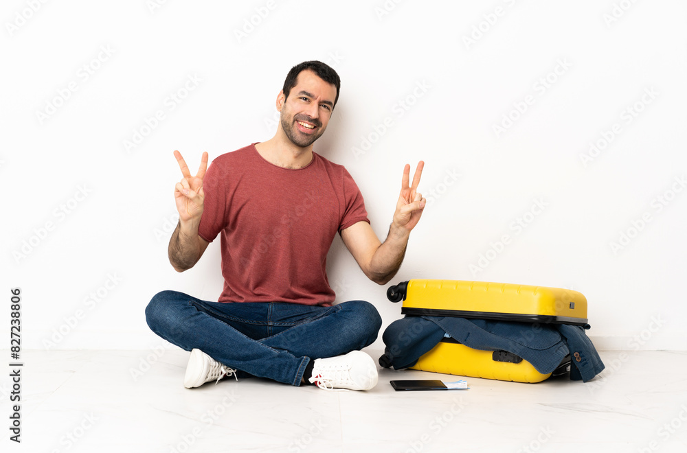 Caucasian handsome man with a suitcase full of clothes sitting on the floor at indoors showing victory sign with both hands Caucasian handsome man with a suitcase full of clothes sitting on the floor at indoors showing victory sign with both hands