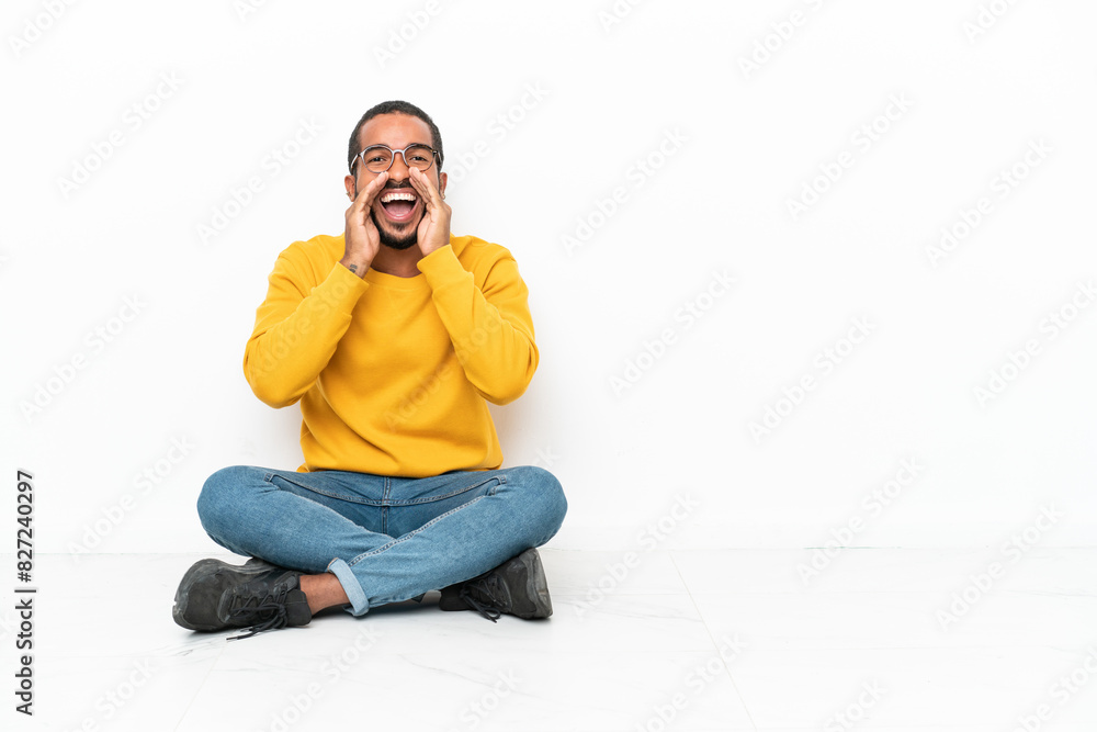 Young Ecuadorian man sitting on the floor isolated on white wall shouting and announcing something Young Ecuadorian man sitting on the floor isolated on white wall shouting and announcing something