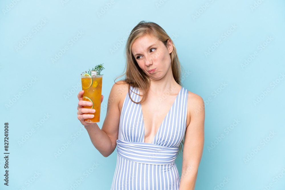 Young blonde woman wearing swimsuit holding a cocktail isolated on blue background with sad expression Young blonde woman wearing swimsuit holding a cocktail isolated on blue background with sad expression