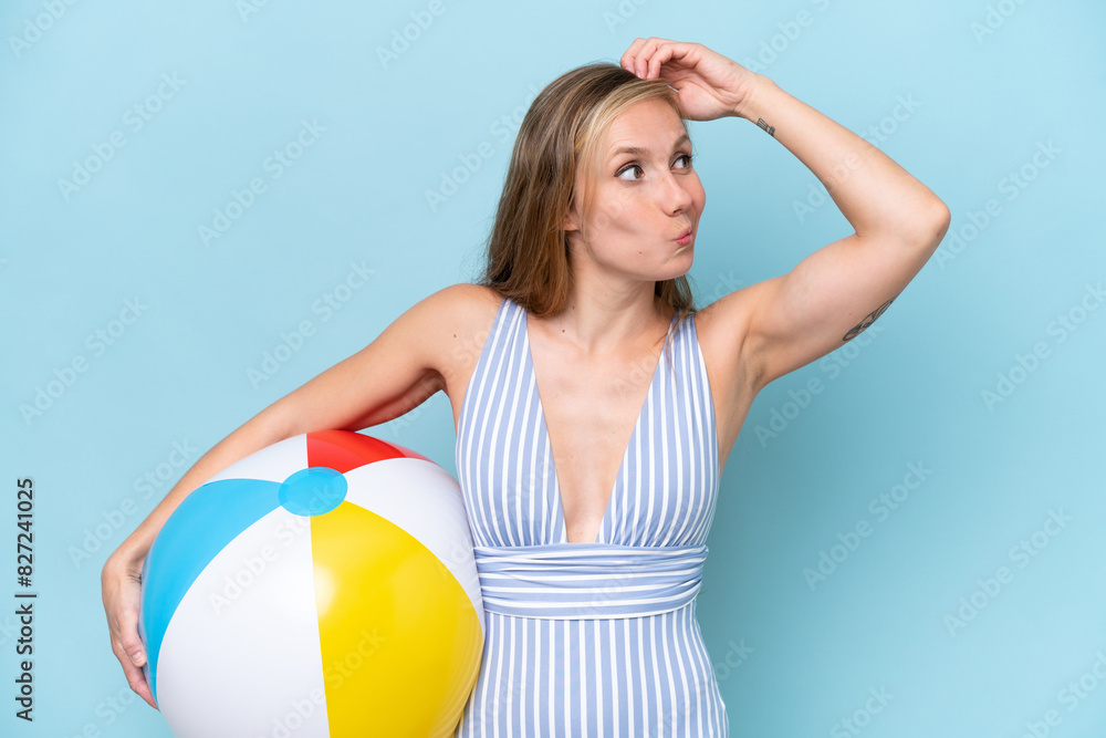 Young blonde woman in swimsuit holding beach ball isolated on blue background having doubts and with confuse face expression Young blonde woman in swimsuit holding beach ball isolated on blue background having doubts and with confuse face expression