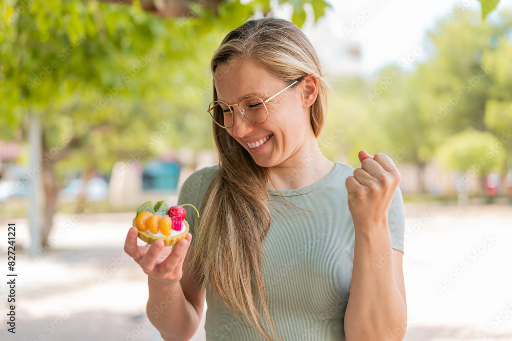 Young blonde woman holding a tartlet at outdoors celebrating a victory Young blonde woman holding a tartlet at outdoors celebrating a victory
