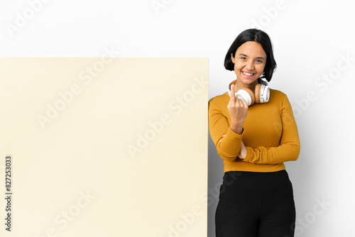 Young woman listening music with a big empty placard over isolated background doing coming gesture photo