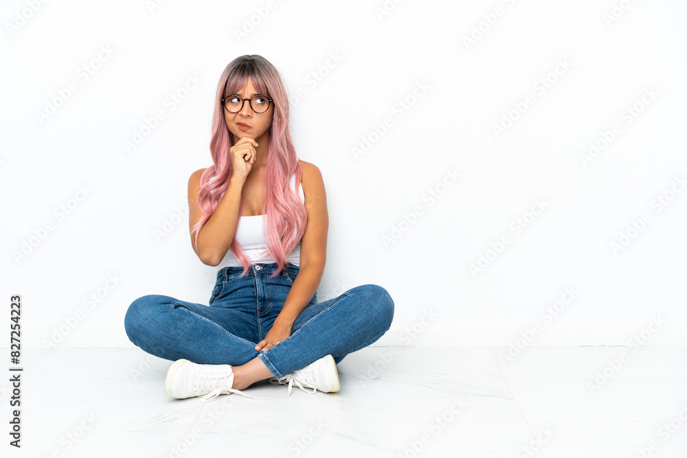 Young mixed race woman with pink hair sitting on the floor isolated on white background having doubts and with confuse face expression Young mixed race woman with pink hair sitting on the floor isolated on white background having doubts and with confuse face expression
