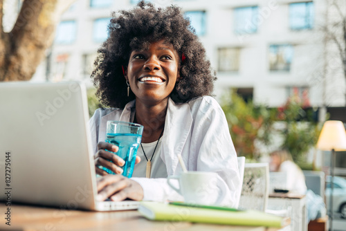 Beautiful black female student sitting at the coffee bar table and using her laptop computer for work and learning. Bring sunny day. Old city buildings in the background.