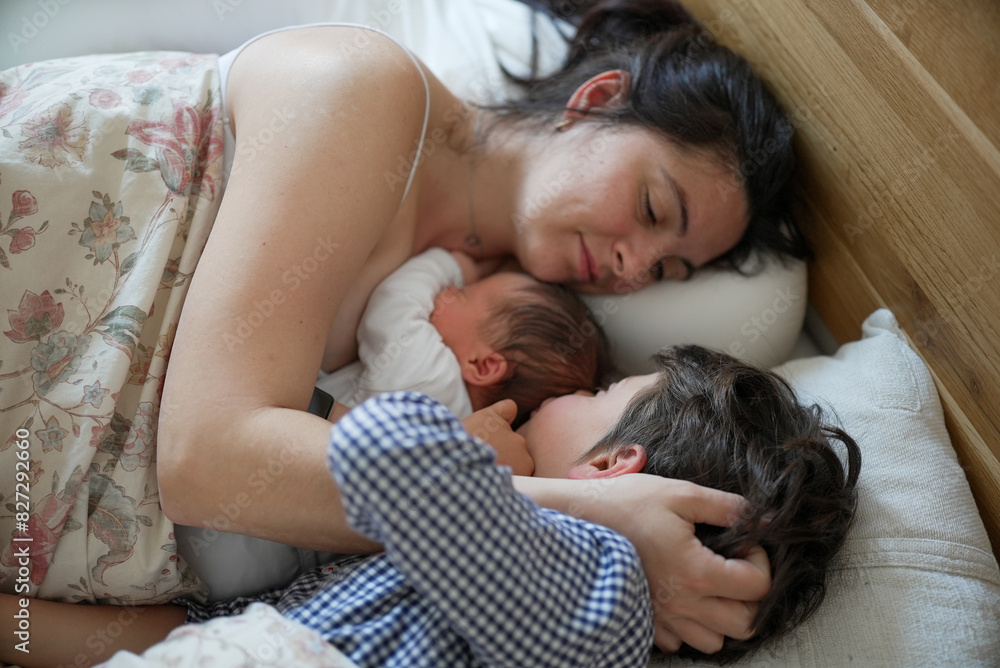 Mother cuddling newborn baby and older son in bed, sharing a loving family moment. The scene captures maternal love, warmth, and the bond between mother and children Mother cuddling newborn baby and older son in bed, sharing a loving family moment. The scene captures maternal love, warmth, and the bond between mother and children