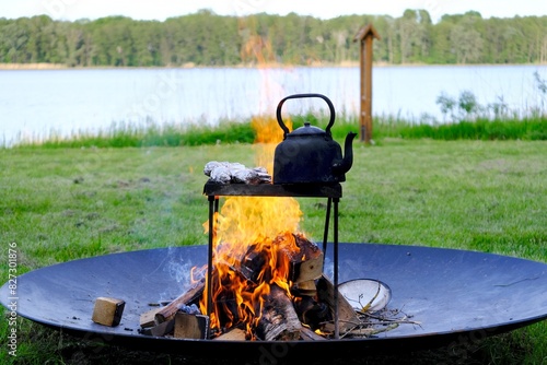 An old metal kettle stands among the flames of the fire. There's grilled food in aluminum foil. Picnic by the lake