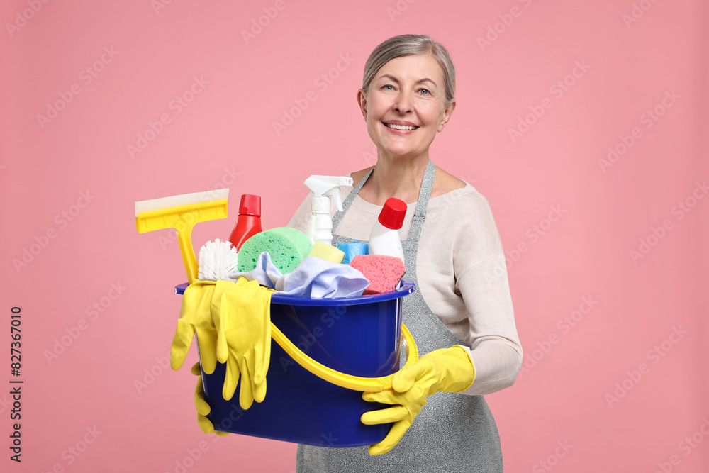 Happy housewife holding bucket with cleaning supplies on pink background Happy housewife holding bucket with cleaning supplies on pink background