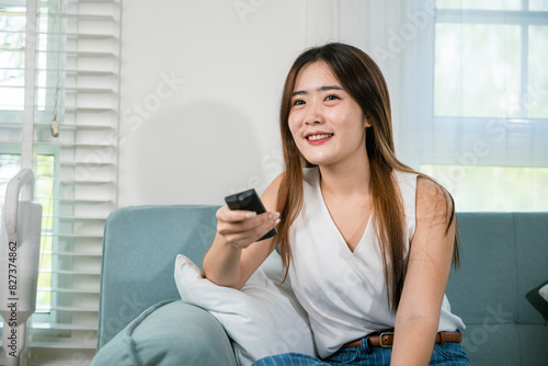 Asian young woman smiling sitting relax watch TV holding remote control on sofa in living room, Happy female fun movie holding remote watching television, Activity lifestyles concept