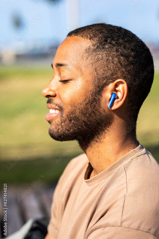 Fototapeta premium A joyful blind African young man basks in the sun at a park near the beach on a bright sunny day. He listens to music with headphones and happily responds to messages on his mobile phone.