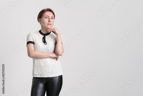 Portrait of serious businesswoman looking at camera with one hand raised to chin. Business person wearing white short sleeve blouse and black leather tight trousers, standing on white background