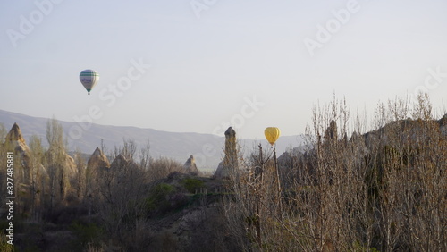 Two hot air balloons flying over the grassy landscape of Cappadocia, Turkey, with iconic fairy chimney rock formations in view. The scene captures the region’s unique natural beauty and the popular 