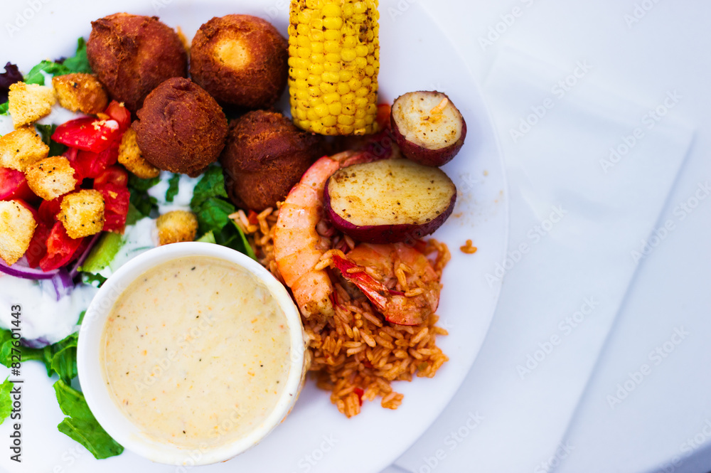 closeup of a seafood boil with corn and shrimp, hushpuppies, brown rice and a salad with dressing on a white plate on a white tablecloth
