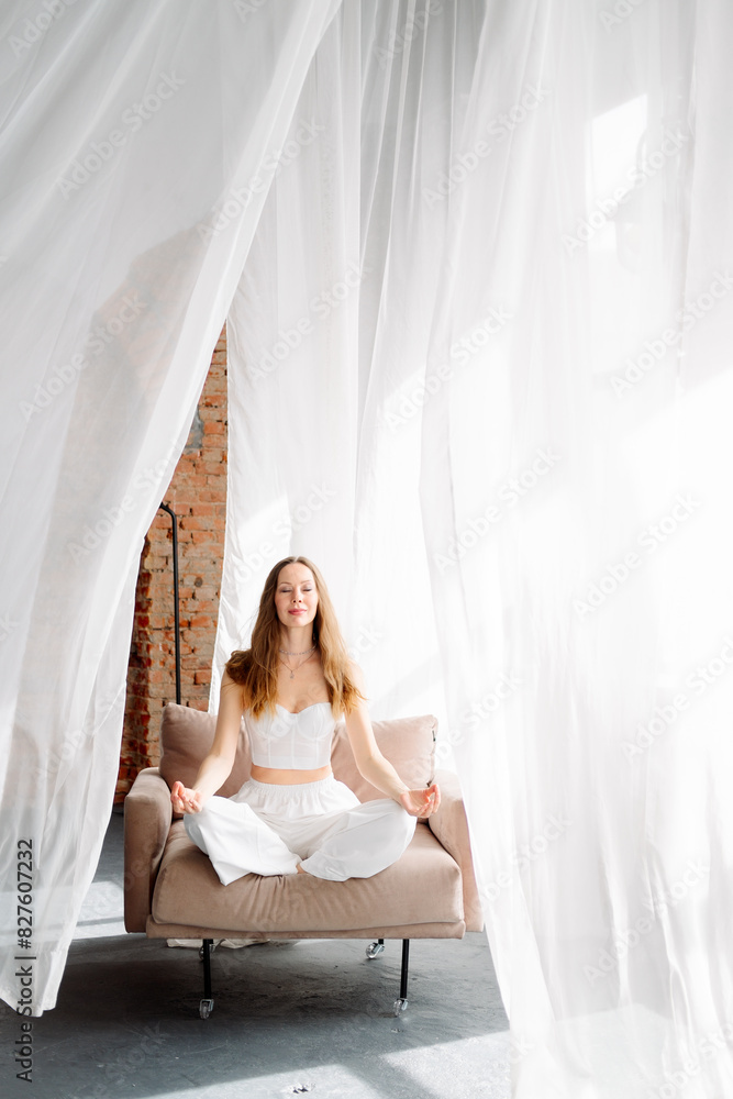 A young female yogi in white clothes sits in the lotus position on a chair against a background of white and airy curtains in a loft studio. A young female yogi in white clothes sits in the lotus position on a chair against a background of white and airy curtains in a loft studio.