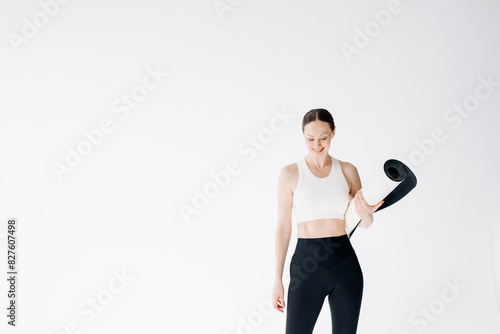 Beautiful and slender woman in a short sports top stands with a mat in her hands, on a white background, copy space
