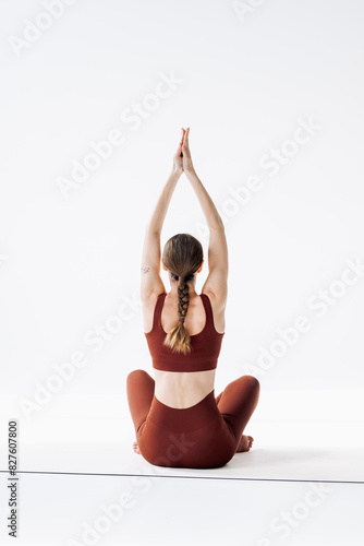 A slender woman in a brown sports uniform with a pigtail sits in an asana on a white background and meditates, rear view. Yoga classes in the studio