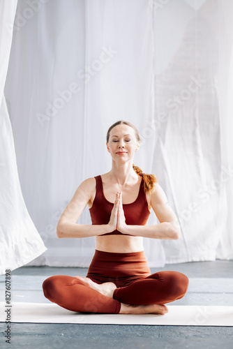 A slender woman in a brown sports uniform sits in the lotus position, with her eyes closed, against the background of white curtains and meditates. Yoga classes in the loft studio
