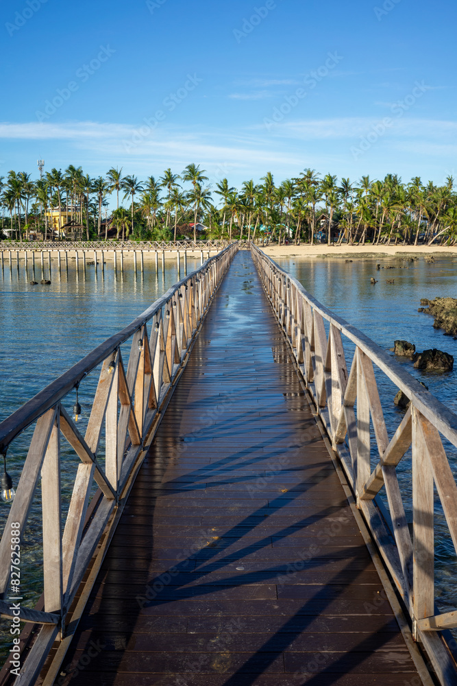 Wooden boardwalk leading to a lush, palm-covered island at Cloud 9, Philippines. The clear blue sky and surrounding waters enhance the tropical beauty of this picturesque location.