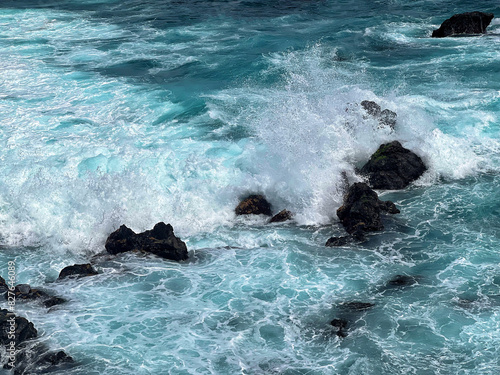 Aerial photo of a sea water surface. Turquoise water with foam on waves - view from above. Water washes black volcanic stones. Stylized background abstract texture photo. Tenerife, Puerto de la Cruz