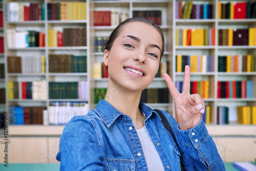 Selfie portrait of high school student girl showing hand gesture for victory  inside library