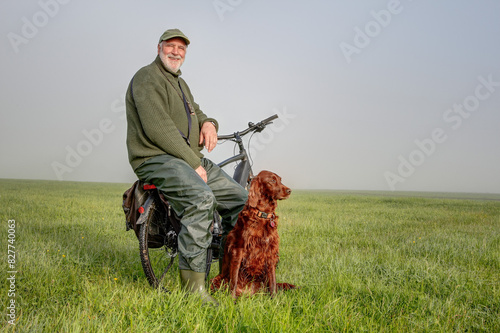 An elderly cyclist pauses in the early morning on a green meadow. He sits on the luggage rack, his Irish Setter in front of him. They both watch the landscape as the sun slowly breaks through the fog.