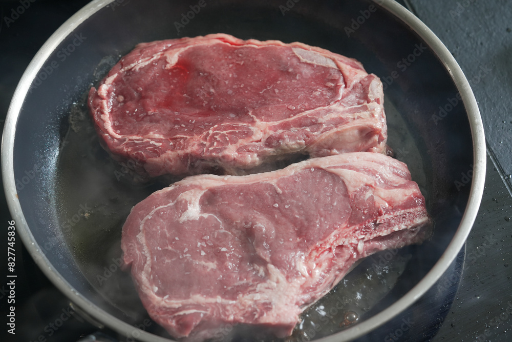 Two raw ribeye steaks searing in a hot pan, emitting steam, capturing the initial stages of cooking, suitable for culinary food preparation, and steak recipe content Two raw ribeye steaks searing in a hot pan, emitting steam, capturing the initial stages of cooking, suitable for culinary food preparation, and steak recipe content