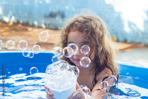 Uma criança dentro de uma piscina infantil azul, brincando, com bolhas de sabão ao redor, no quintal de casa. photo