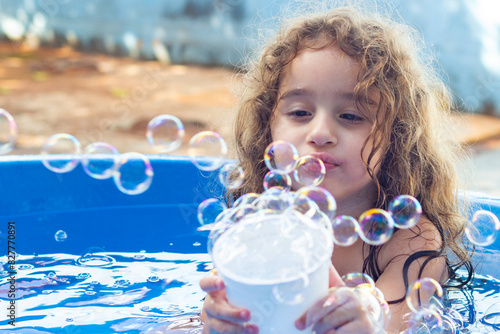 Uma garotinha dentro de uma piscina infantil azul, brincando com bolhas de sabão no quintal de casa. photo