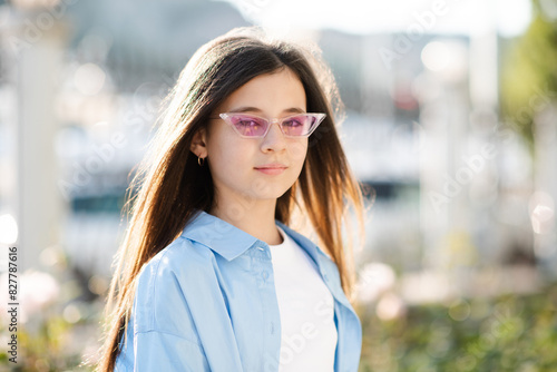 Stylish teenage girl 10 - 12 year old wearing sun glasses and blue shirt posing over nature sunny background. Looking at camera. Summer vacation season.