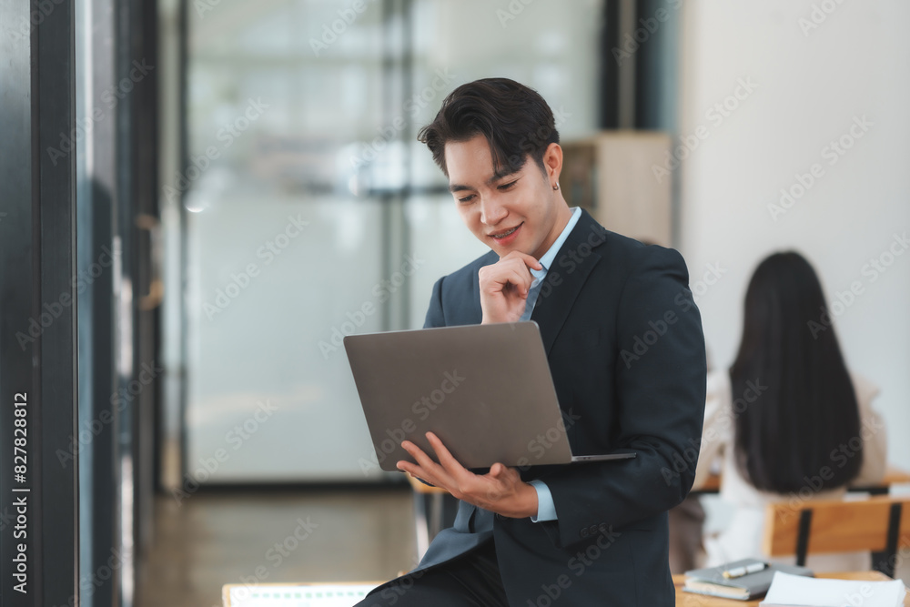 A confident businessman is sitting and working on his laptop in a modern office setting, focused on his work with a thoughtful expression