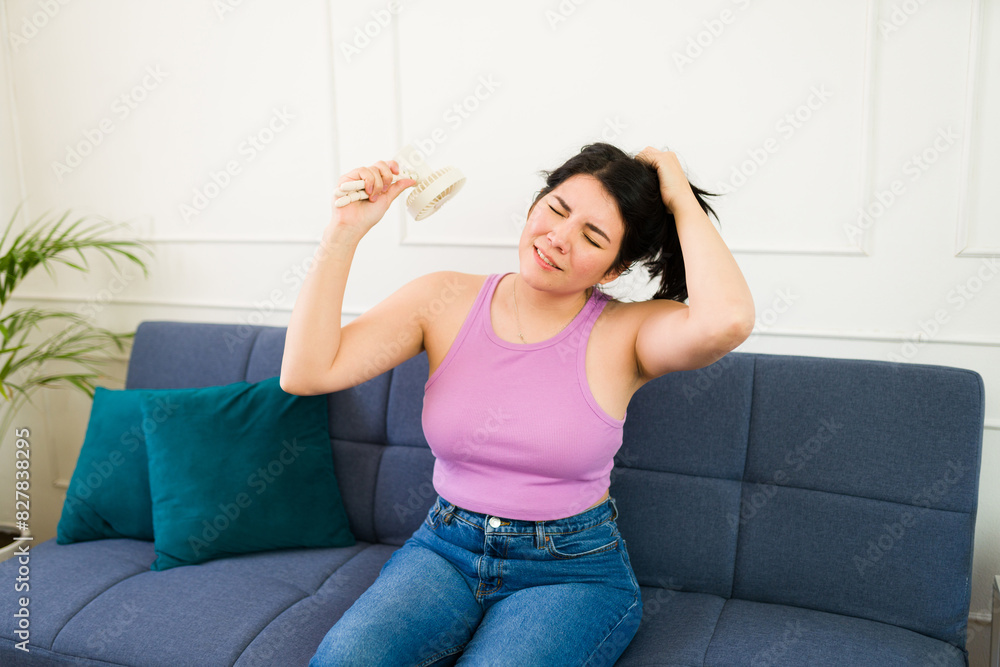 Young woman at home using a hand fan to cool down and beat the summer heat, visibly sweating Young woman at home using a hand fan to cool down and beat the summer heat, visibly sweating