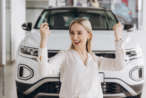 Cheerful woman standing near vehicle in dealership center and raising hands with excitement, copy space. Happy owner. Overjoyed Caucasian lady holding keys of new car after buying.